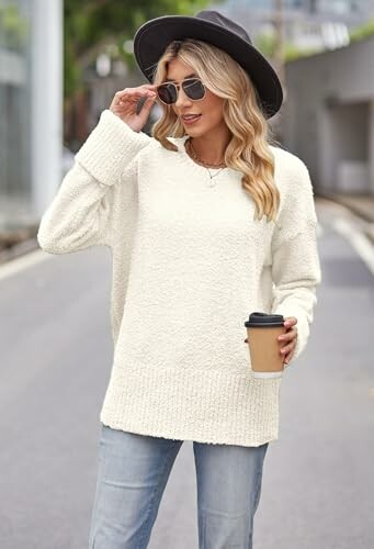 Mujer con suéter blanco, sombrero negro y gafas de sol, sosteniendo un café en la calle.