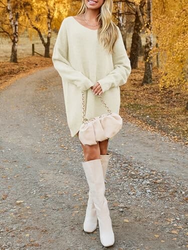 Mujer con suéter beige y botas largas en un camino otoñal.